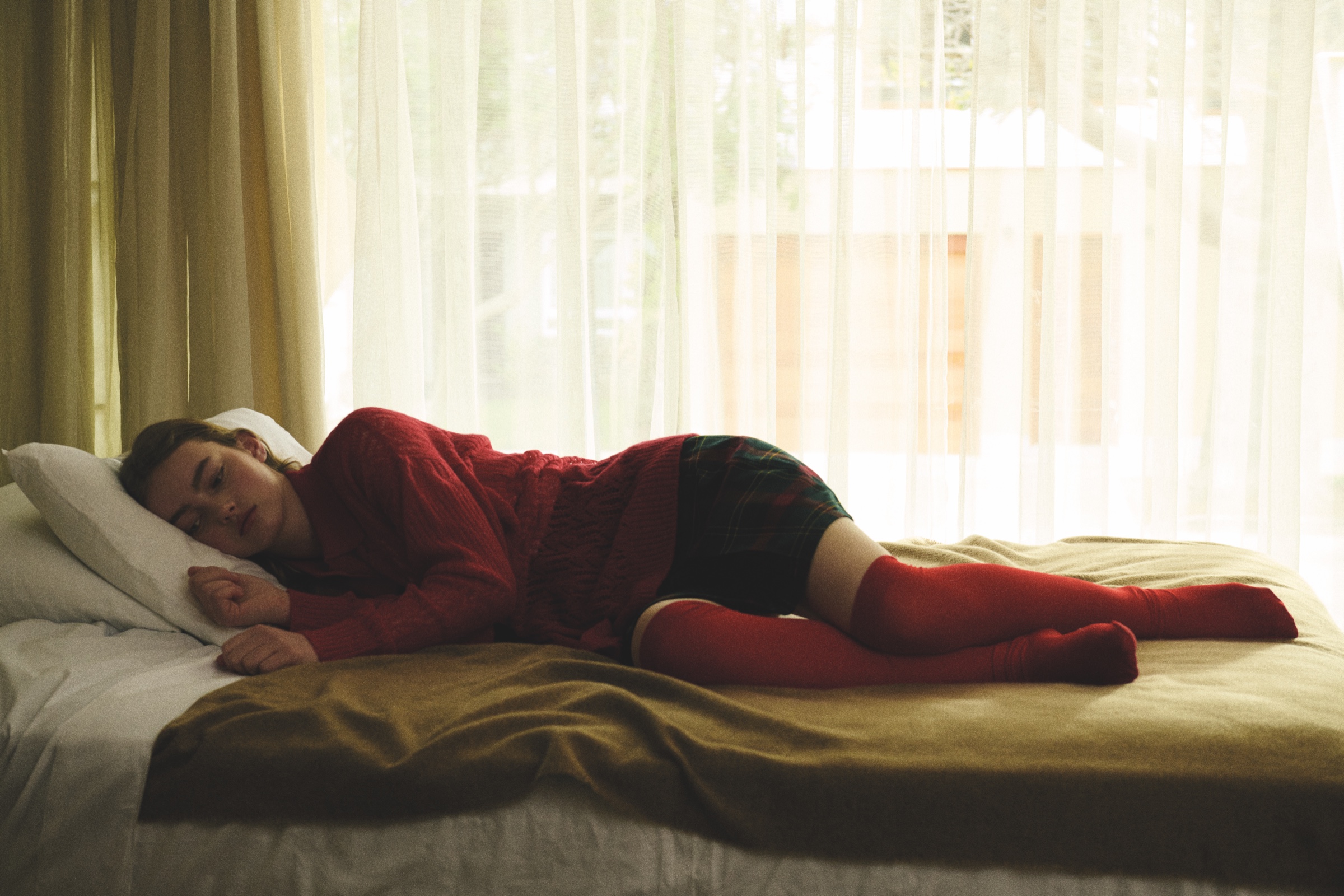 A student in a red sweater lies on a sunlit bed.